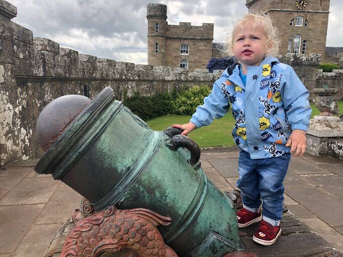 A young boy stands next to a cannon at Culzean Castle, resting one hand upon it. The clocktower is in the background.