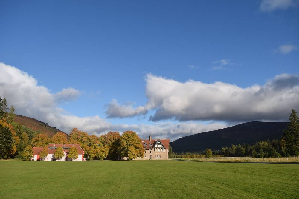 A view looking across a vast expanse of lawn to Mar Lodge on a bright sunny day. The trees are green and beginning to turn into their autumn colours in some places. Tall mountains can be seen in the background.