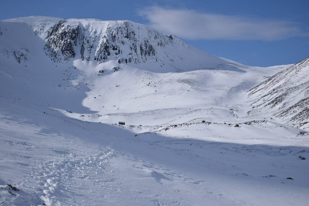 A mountain landscape in winter, covered in thick snow. A trail of footprints lead across the glen towards the summit.