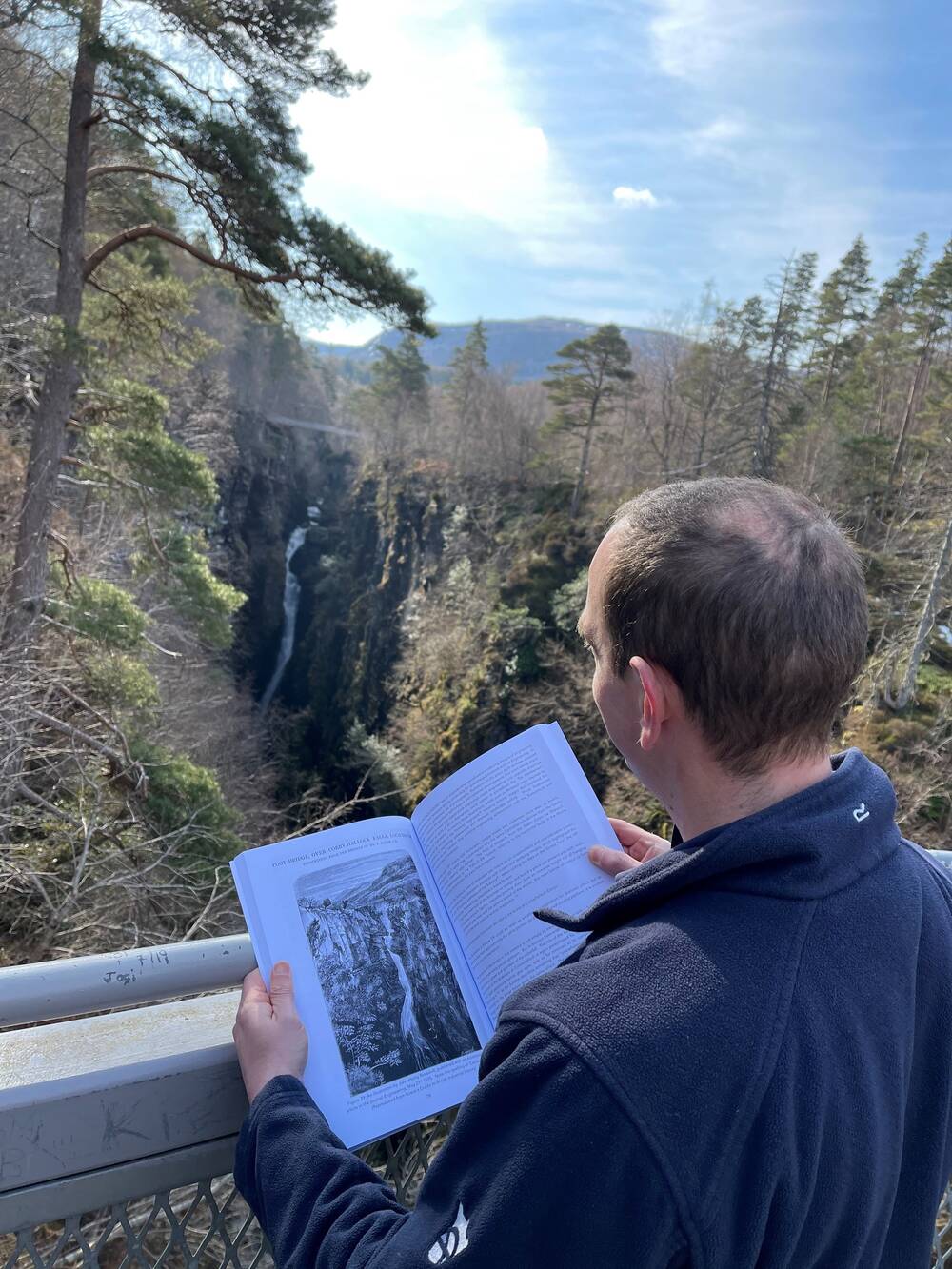 A man stands with his back to the camera, holding an open book that rests against railings. He is looking at a view of a suspension bridge that crosses a deep gorge. The same view is illustrated on the open page of the book.