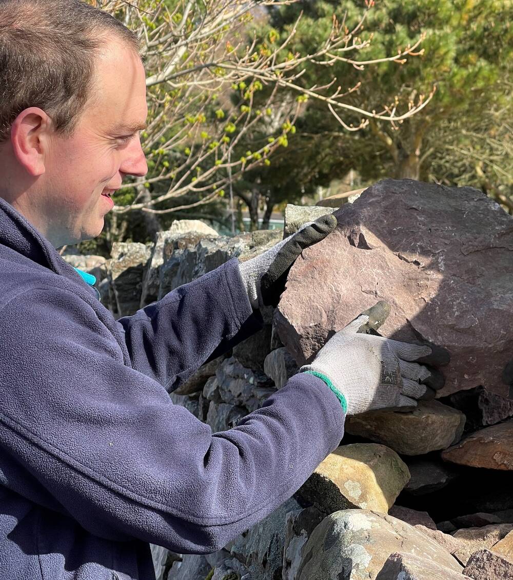 A smiling man puts a large stone into the correct place on top of a drystone wall. He is wearing a navy fleece and grey work gloves.