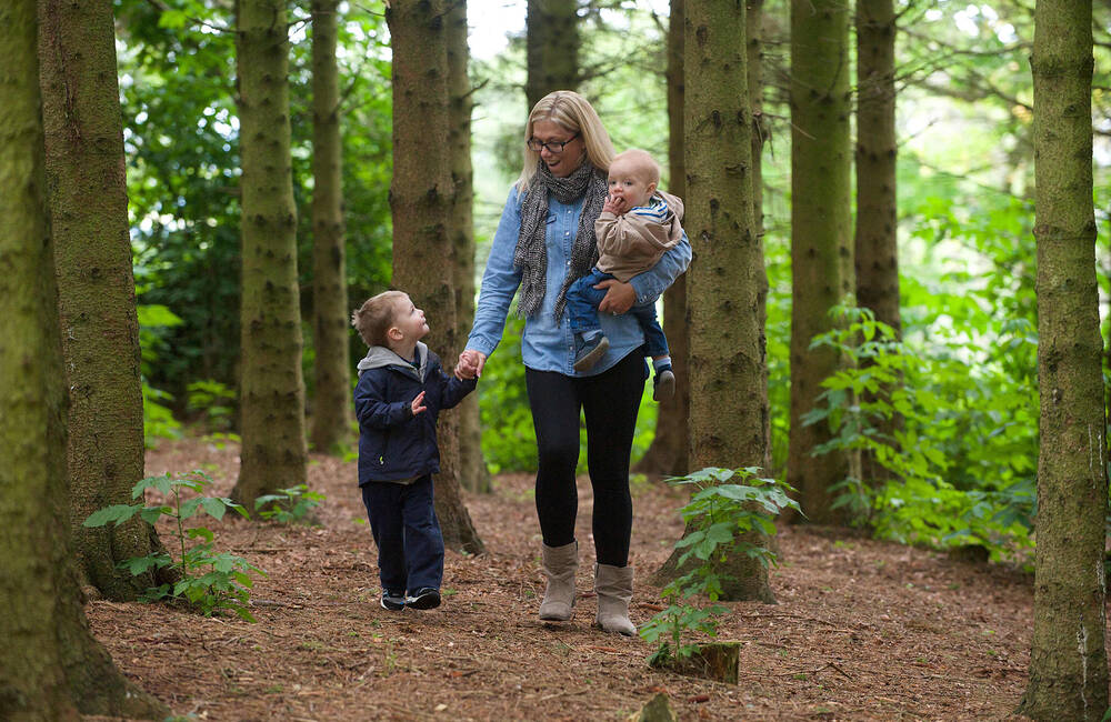 A mum walks along a woodland path, carrying a toddler in her arms and holding the hand of a young boy next to her.