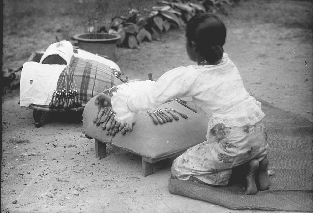 A glass plate negative, black and white image of a back view of a girl kneeling on a blanket while leaning against a weaving device.