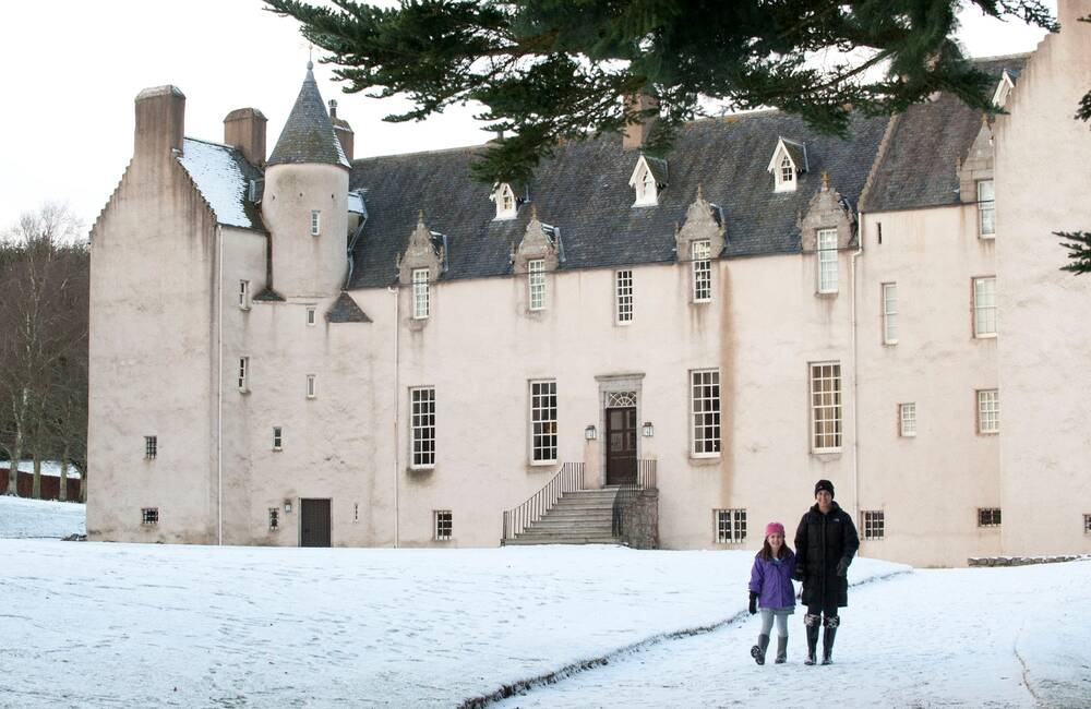 A view of the long Victorian wing of Drum Castle in winter, with snow covering the ground. A woman and child walk handing hands along the snow-covered path outside.