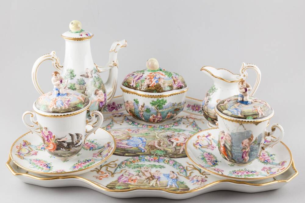 An extremely expensive-looking porcelain coffee set is displayed against a plain grey background. On a shaped tray sits a pair of two-handled cups with covers and saucers, a coffee pot, milk jug and a quatrefoil sugar bowl. The design features flowers and classical figures.