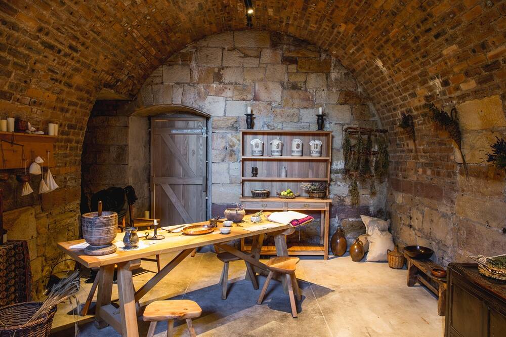 A room with curved brick walls and ceiling set up like a traditional apothecary, with a long wooden table and shelves with pots and jars of ingredients for remedies.