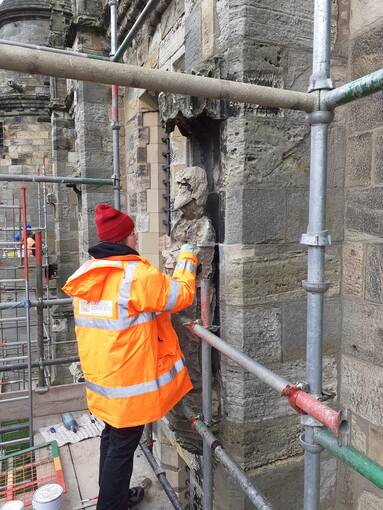 A woman in an orange hi-vis jacket and red woolly hat applies a resin to a stone statue set in a niche on a palace wall. She is standing on a scaffolding platform.