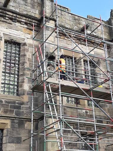 A view of the exterior wall of Falkland Palace with scaffolding against one section. A woman in an orange hi-vis jackets stands on a high platform, working on a stone statue set in a niche on the wall.