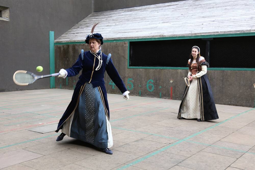 Two women in historical dresses hold rackets and play a game of real tennis on the royal tennis court.