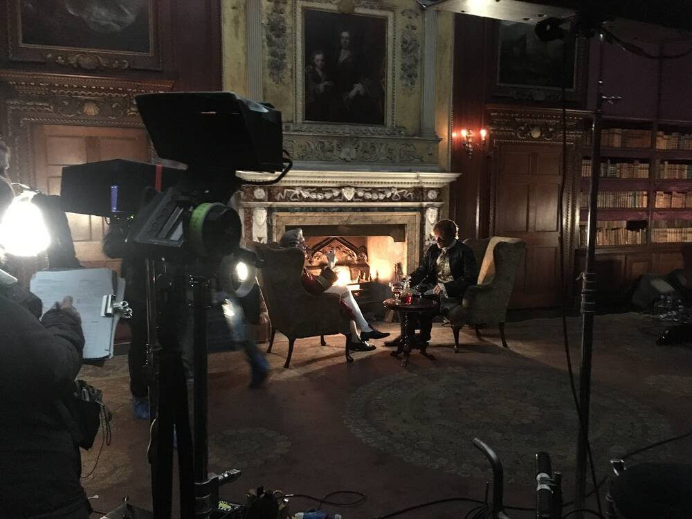 Two actors are filmed in conversation, sitting in chairs in front of a fireplace in a grand drawing room of a stately home. The camera stands and lights can be seen in the foreground, along with a person holding a clipboard.