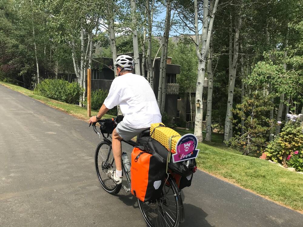 A man rides a mountain bike with fully packed paniers and a little omega-shaped sign at the back, reading National Trust for Scotland Love. He is riding along a tarmac road beside some houses.
