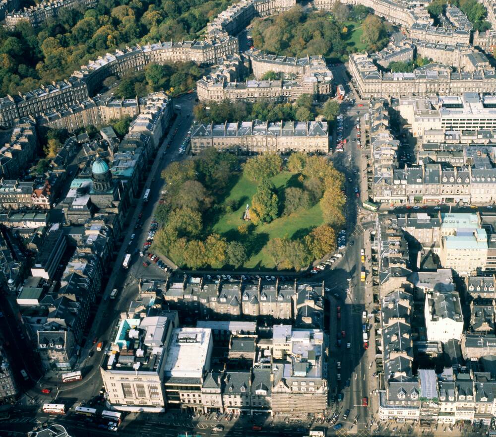 The streets of Edinburgh's New Town viewed from the air, with Charlotte Square at the centre. A road wraps around a green space with trees, while houses line the sides.