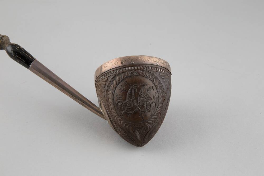 A close-up of the bowl end of a ladle shaped from a coconut shell. The shell has leafy carvings upon it, and has a silver rim with words etched upon it. The silver join to the wooden handle rests to the left.