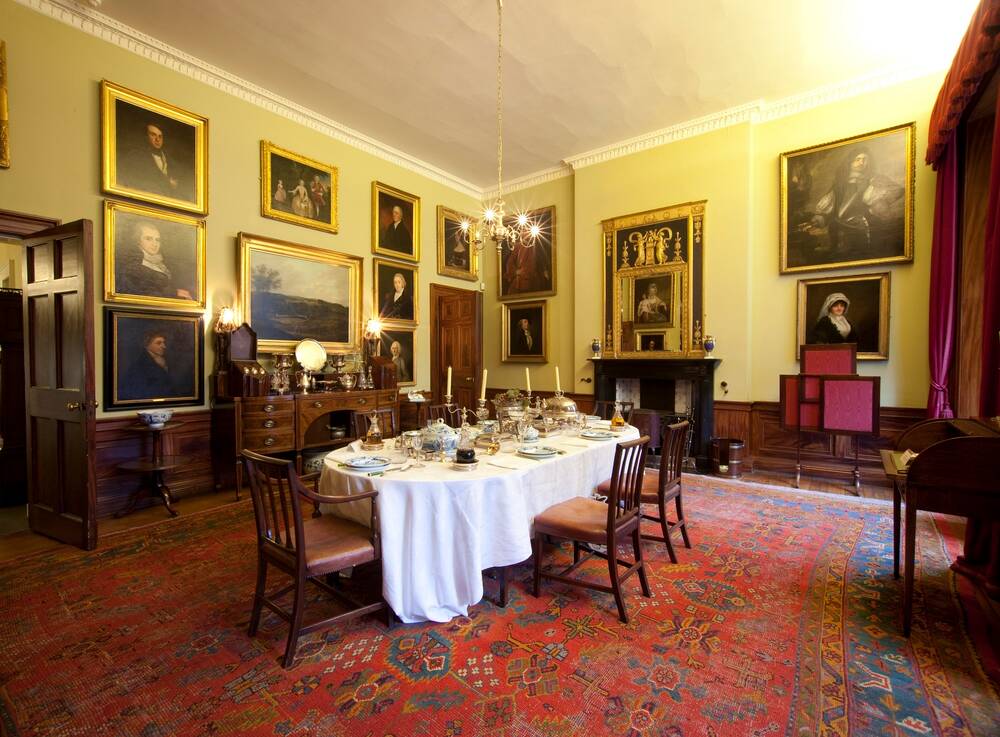 A dining room in a Georgian House, showing the table laid in the centre of the room. The walls are covered by gilt-framed portraits. Red curtains hang at the windows and the floor is covered with a patterned red carpet.