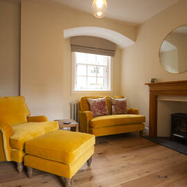 Interior cream room with two yellow armchairs and a yellow footstool. There is a fireplace with wooden mantlepiece, a mirror hangs above the fireplace. 