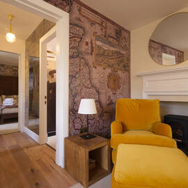 A single yellow armchair and footstool sit in front of a fireplace with a white wooden mantlepiece. One wall is papered with a historic map of Scotland.