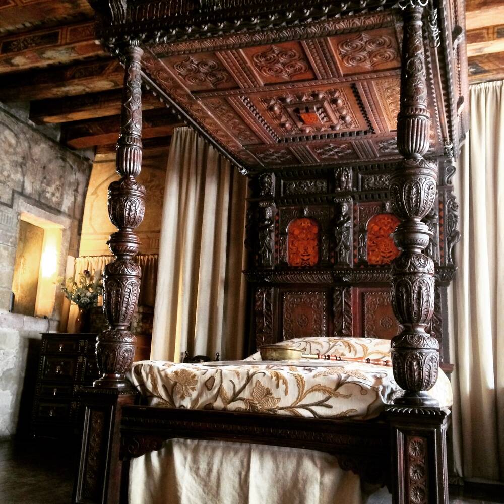 An elaborately carved, wooden four-poster bed stands in the middle of a wood-panelled room. It is covered with an embroidered blanket. A long-handled bed pan lies on top.