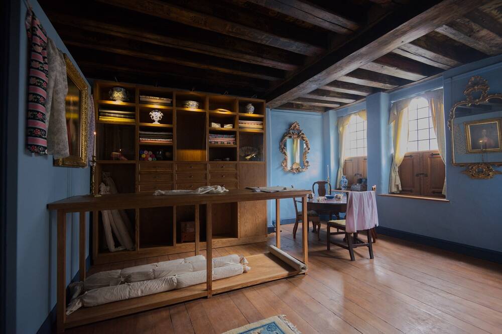 A view of a re-created 18th-century draper's shop. A large work bench stands before a large wooden shelving cabinet. The walls are painted blue, and gilt-framed mirrors are hung around the room.