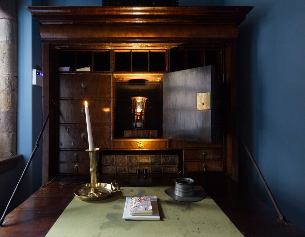 A view of a dark wooden cabinet. The door in the middle is open, to reveal an oil lamp inside. The cabinet holds lots of different sized drawers. A table stands before it, with a lit candle on it.