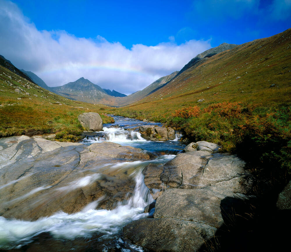 A rainbow arches over the pyramid-shaped Goat Fell, with a rushing stream in the foreground.