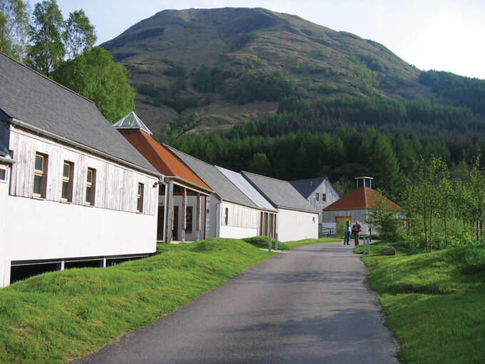 A row of single-storey buildings stand beside a path. The buildings are wooden and stand on stilts. A tall mountain looms in the background.