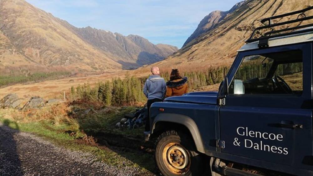 A couple sit on a wall looking down Glencoe. They are sitting just in front of a navy Land Rover, that has Glencoe & Dalness written on the passenger door.