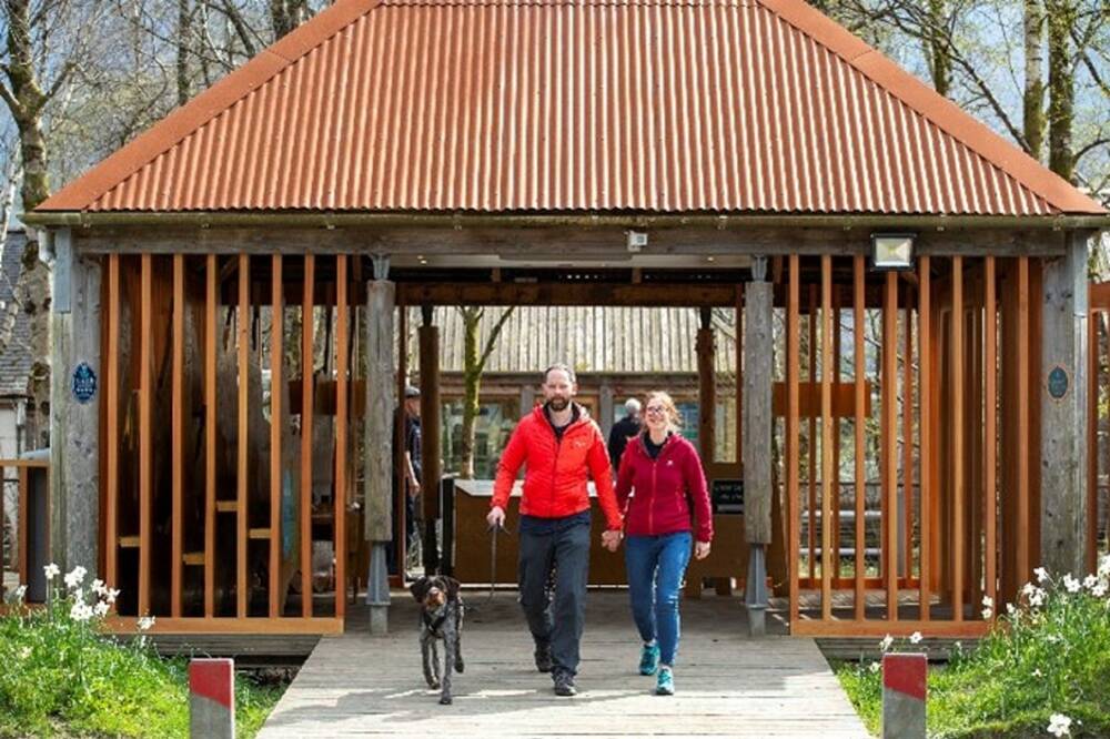 A man and a woman, both wearing red outdoor jackets, walk hand in hand along a wide boardwalk. They're heading away from a covered porch-like area in front of a visitor centre.
