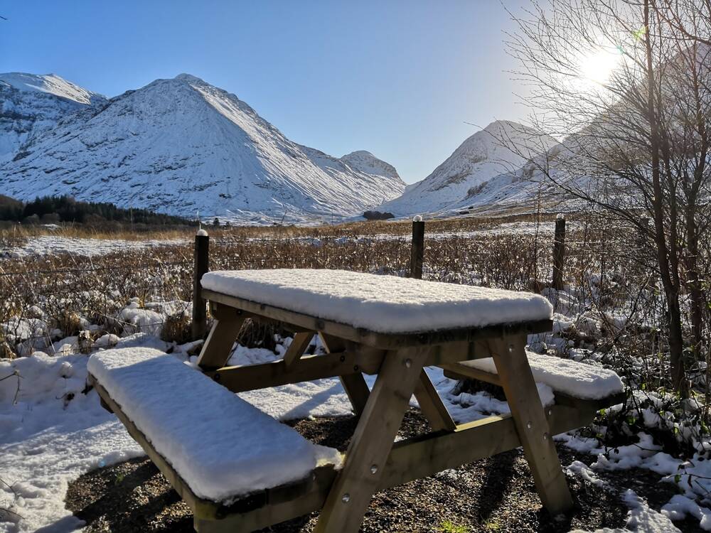 A picnic bench covered with a thick layer of snow stands in the foreground, with the dramatic snow-covered mountains of Glencoe rising in the background. The sun is low and gleams through a gap between the hills.