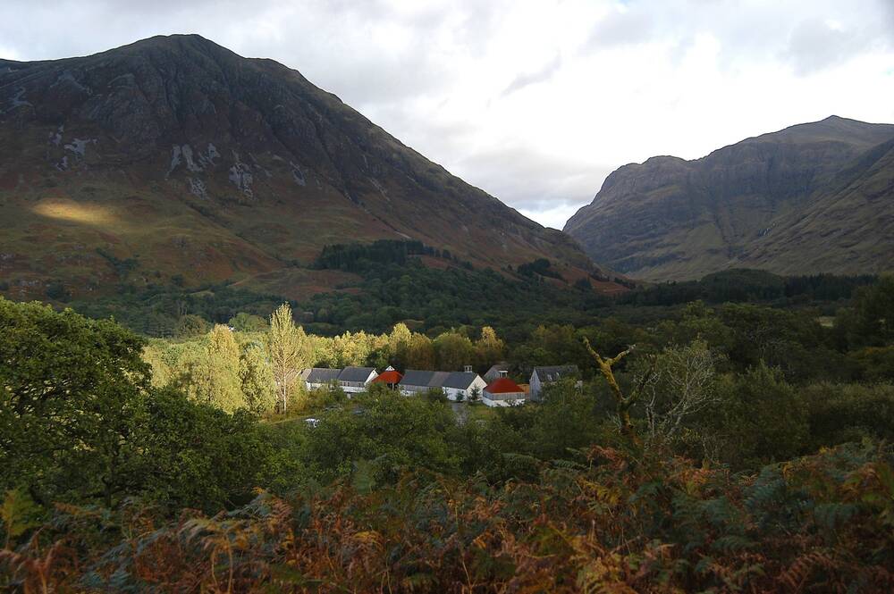 Glencoe Visitor Centre and related buildings form a cluster, surrounded by woodland and nestling in the glen. Tall mountains loom in the background.