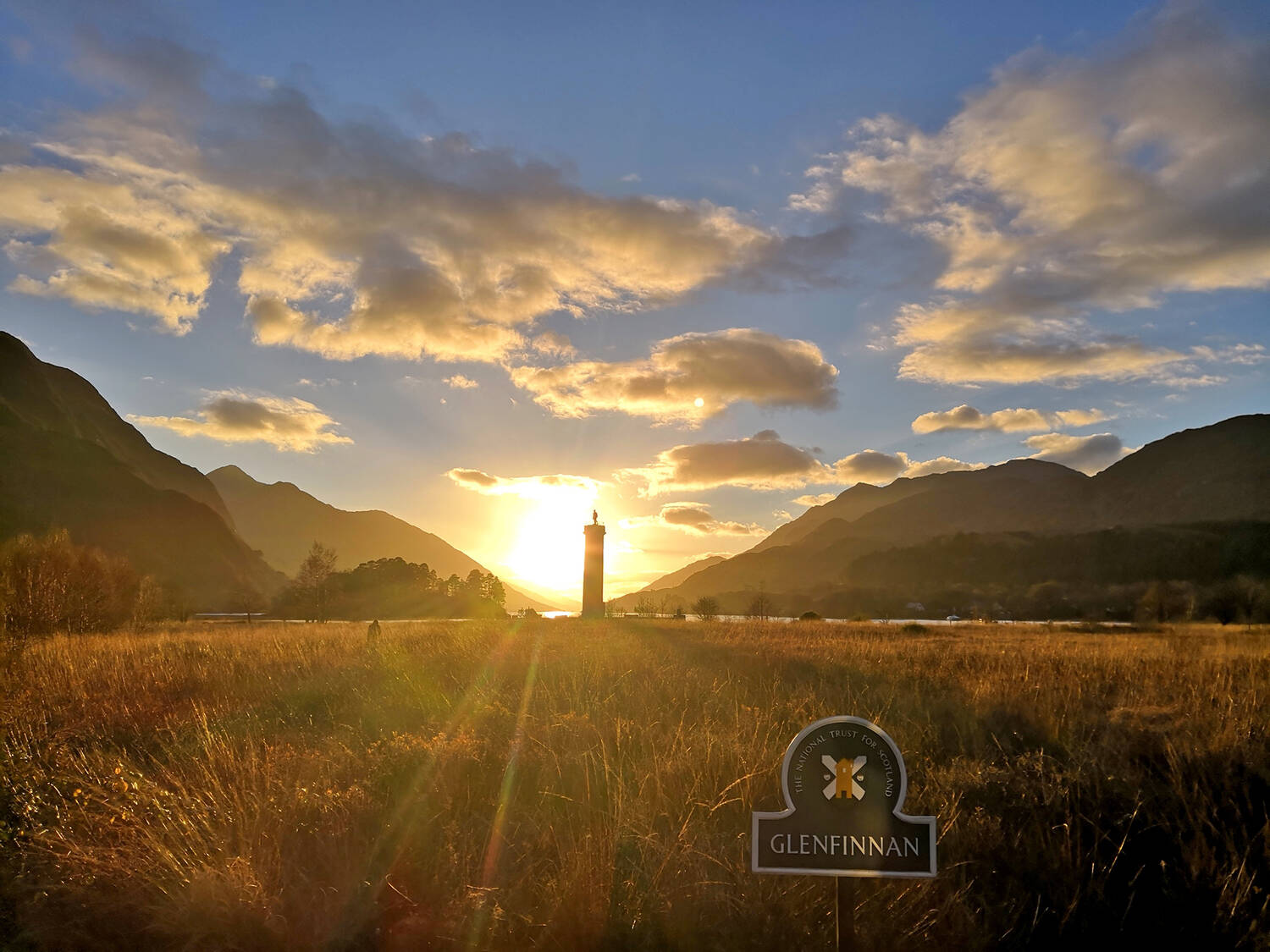 The sun sets over a loch, centred in a narrowing glen. Silhouetted against the sunset is a tall, thin column with a statue on top. A sign reading Glenfinnan stands in the field in the foreground.