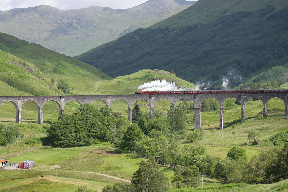 A red steam train puffs across a long stone viaduct, with tall mountains rising from a green glen in the background.