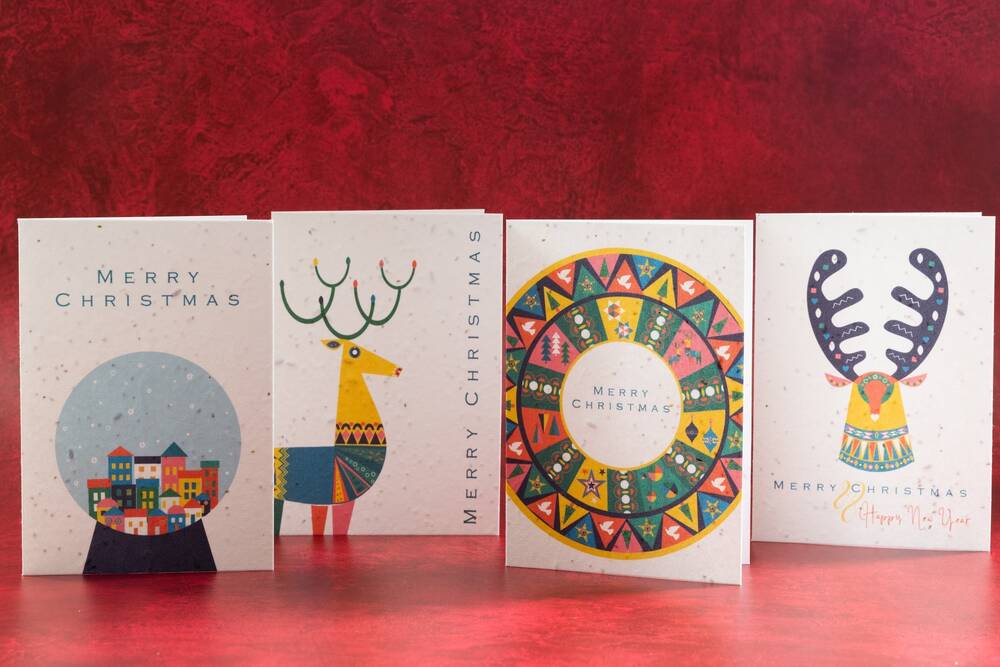 Four Christmas cards, printed on seeded paper, stand against a red marbled background. From left to right, they feature a snow globe scene, a reindeer, a wreath and a stag's head.