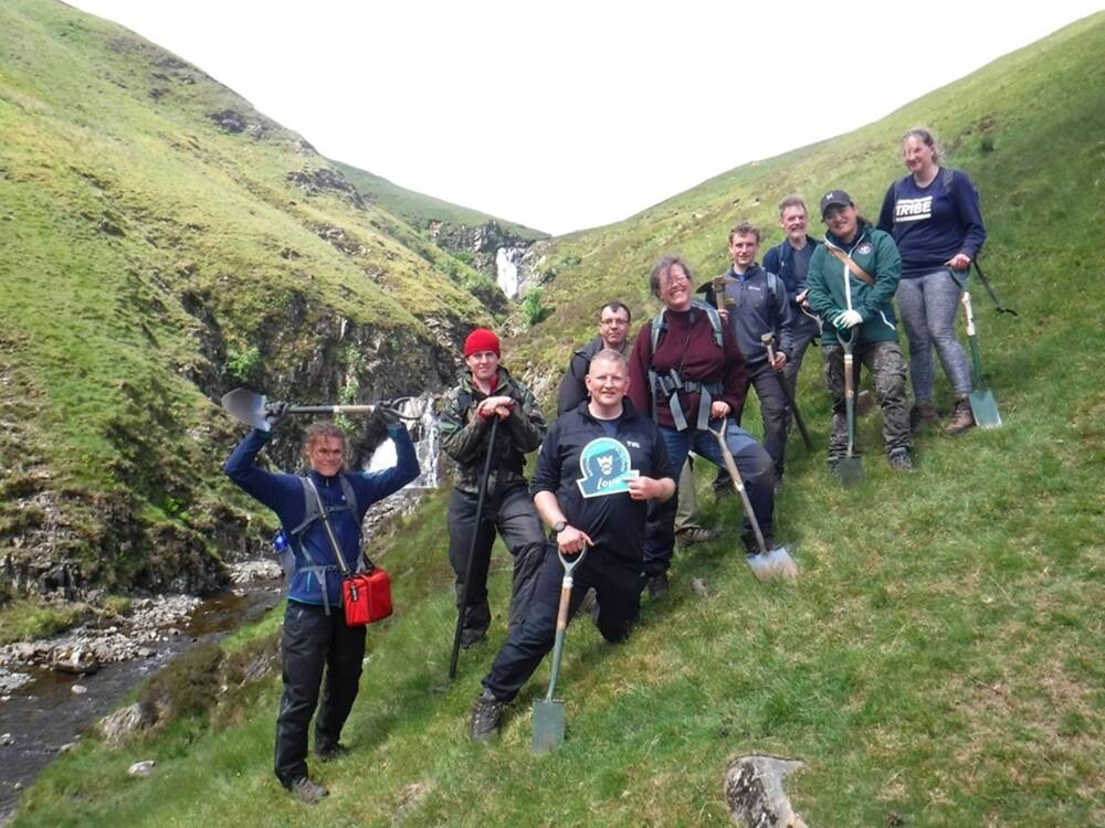 A large group of people stand on the mountainside beside a waterfall. They all are holding spades, or similar tools. The lady to the far left hold her spade above her head. The man in the centre holds a National Trust for Scotland ‘Love’ sign.