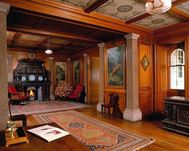 The wood-panelled entrance hall to Haddo House. Rugs lie on the polished wooden floor, a very grand wooden fireplace can be seen on the far wall; and a visitor book lies open on the table in the foreground.
