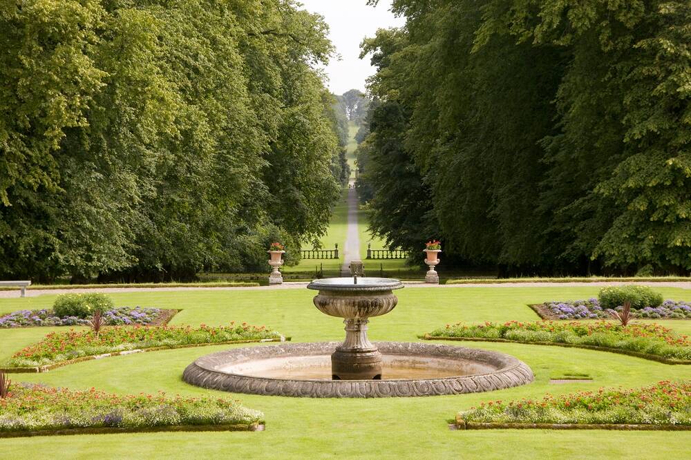 A view of a fountain with a circular surround, situated in some formal gardens. Behind the lawns is a country park, with tall fir trees either side of a long grassy avenue.