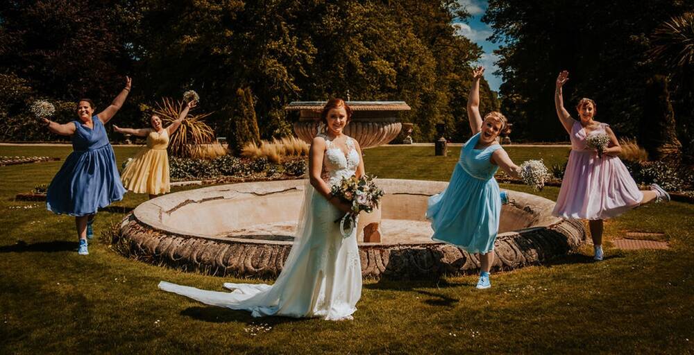 A bride stands in front of a circular fountain in ornate gardens. Behind her and arranged around the fountain are four bridesmaids, all in pastel coloured dresses. They are jumping and holding their bunches of flowers in the air. They are laughing!