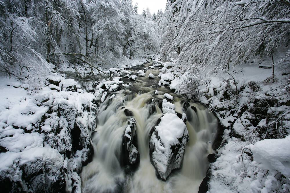 The Hermitage in winter. A waterfall surges over snow-covered rocks, with snow-laden trees hanging over the riverbanks.