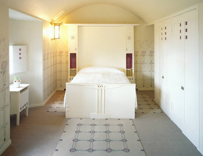 A view of the main white bedroom in the Hill House, with the bed centred. Purple silk hangings are displayed either side of the headboard. A small cabinet stands in the window alcove. A lamp is lit beside the bed.