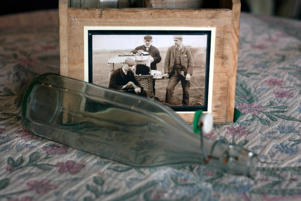 An empty glass bottle lies on its side in front of a wooden box. The box has a sepia photo on the front of three men drinking tea from a flask and holding golf clubs.
