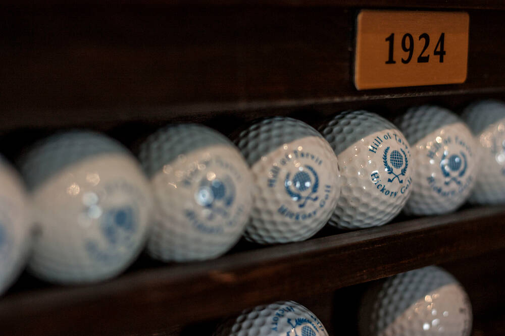 A brown wooden shelf filled with white golfballs, the golfballs have a logo of two crossed golf clubs in blue. Above the shelf is a brass plaque that reads 1924.