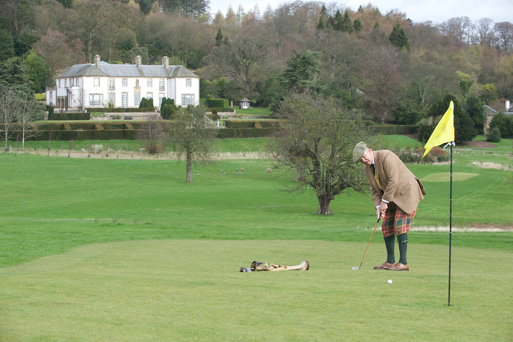 A single golfer stands on the green, next to a yellow flag. The golfer is swinging a golf club, preparing to hit a ball into the hole by the flag. In the background is a white mansion house and woodland.