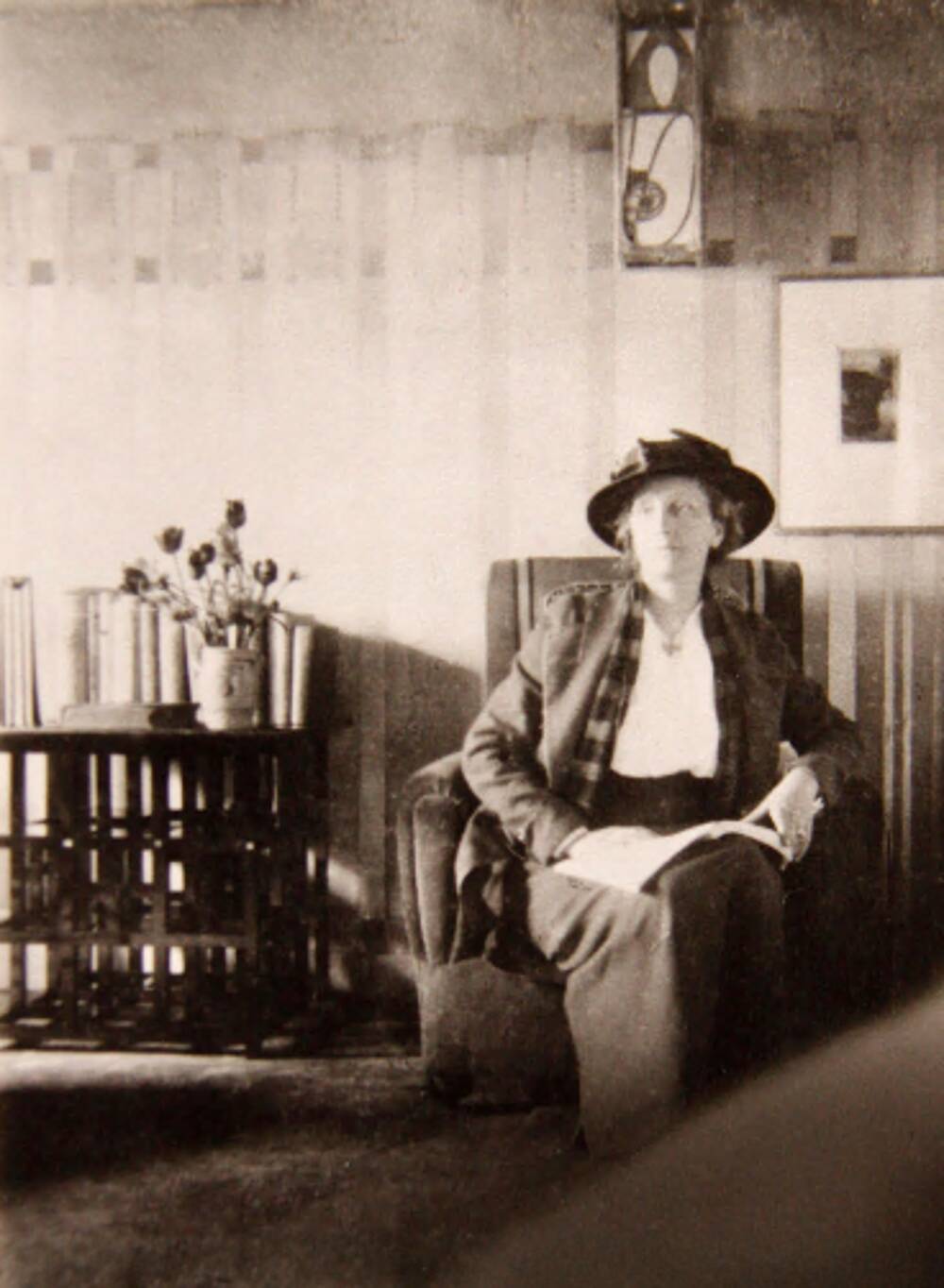 A black and white photo of an Edwardian woman sitting in an easy chair in the Hill House. Mackintosh furniture and wall  decoration surrounds her. She wears a hat, tweed jacket and skirt and has a loose tartan scarf around her neck.