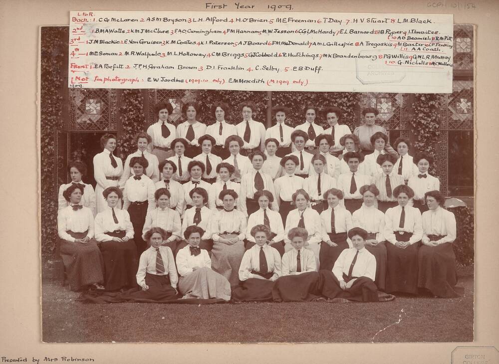 A black and white college photo of rows of young women in the early 20th century. All wear white blouses with a dark narrow tie. At the top of the photo is a handwritten label, detailing the girls' names.