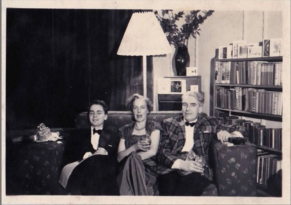A black and white photograph of a woman sitting on a sofa in between an older man and his teenage son. They are smartly dressed and holding drinks. They seem to be in a lounge, with a large standard lamp behind the sofa and a full bookcase to the right.