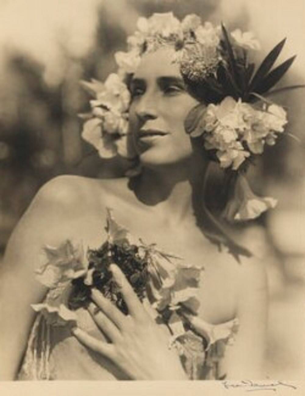 A sepia-tinged photograph of a young woman, holding flowers against her chest. She is wearing a strapless dress and also wears a garland of flowers in her hair.