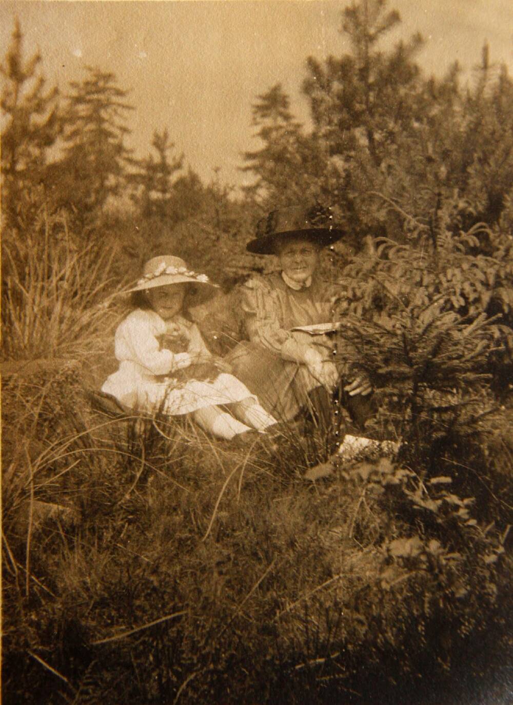 A black and white photo of an older woman sitting next to a young girl on a grassy bank. Trees and shrubs surround them. Both wear wide-brimmed hats to shield their faces from the sun. The woman is wearing a grey or brown dress; the little girl is wearing a bright white dress.