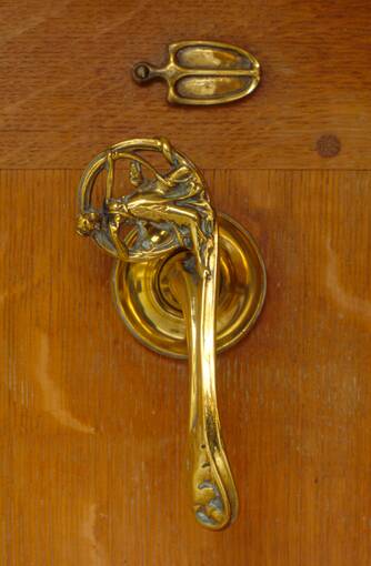 A close-up of a polished metal cupboard handle, with a mini carved figure at the top.