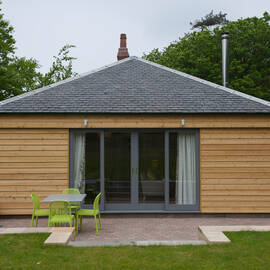 A wooden cabin with a grey tile roof is surrounded by a green lawn and patio area. A garden table and chairs are set out on the patio, in front of the large patio doors. Trees are in the background.