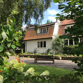 A two-storey cottage with a red tiled roof stands in a pretty garden, with a wooden bench in front. Trees and colourful flower beds frame the image.