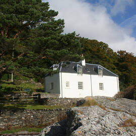 Craggan Cottage, a white two-storey house sits above a rocky shore with trees in the background.